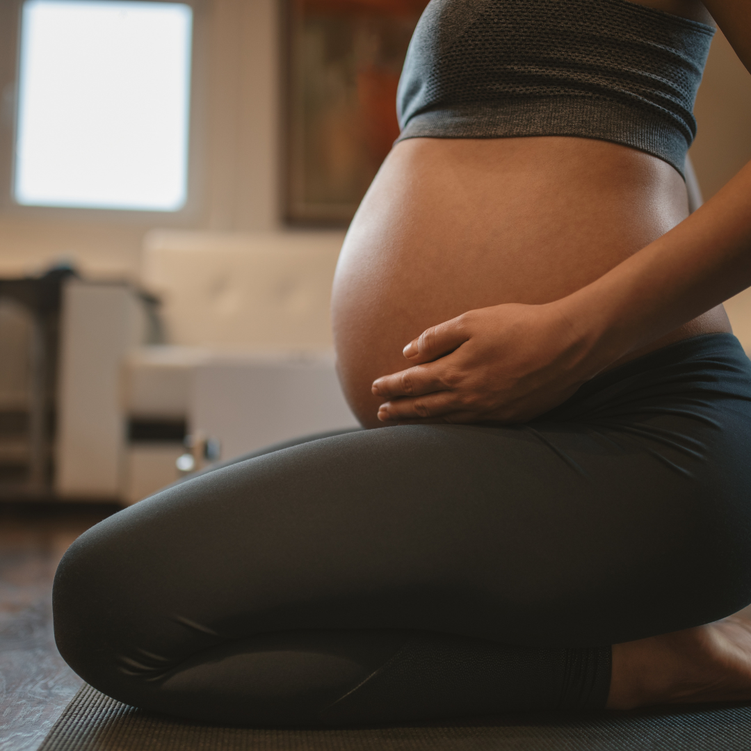 A pregnant woman wearing black leggings and a sports bra kneels on a yoga mat, gently cradling her belly with one hand in a softly lit indoor setting.